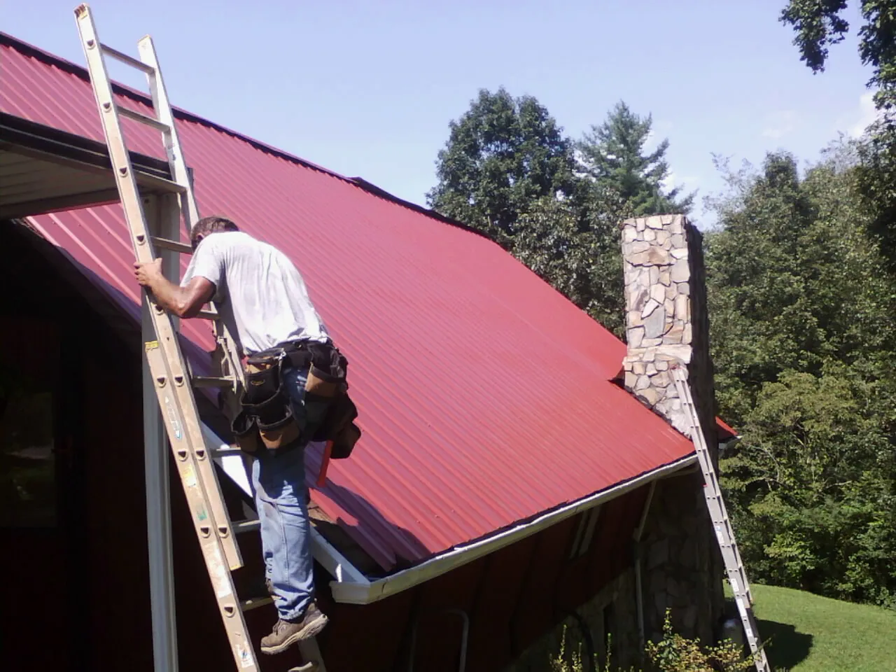 Roofer on a ladder beside a red metal roof and stone chimney — Soffit & Fascia Repair by Roofing Brothers in Green Ridge
